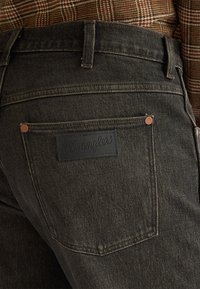 Poche arrière d'un jean en denim noir avec un patch logo Wrangler porté avec une chemise à carreaux marron.