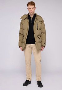 Kurtka puchowa w oliwkowej zieleni z kieszenią na zamek, kapturem i zatrzaskami. Połączona z czarnym swetrem, beżowymi spodniami i czarnymi trampkami.