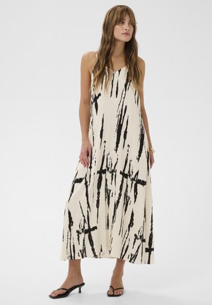 Kobieta w bezrękawowej, luźnej sukience maxi z czarnymi, abstrakcyjnymi pionowymi wzorami pędzla na kremowym tle, w połączeniu z czarnymi sandałami na obcasie.