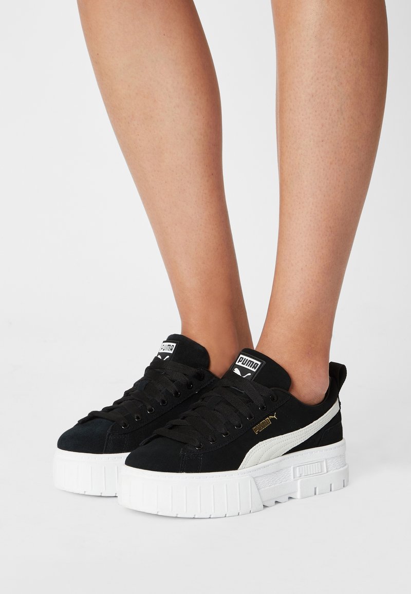 Pieds portant des baskets plateforme Puma noir et blanc avec semelles épaisses sur fond blanc.