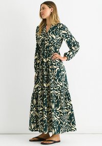 Long green and cream patterned dress with a tiered skirt and button front, featuring long sleeves and an elastic waist. Paired with black sandals.