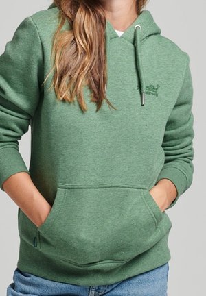 Osoba ubrana w zieloną bluzę z kapturem, z kieszenią z przodu i sznurkami, ręce włożone w kieszeń, w połączeniu z niebieskimi dżinsami.