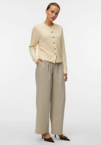 Cardigan a coste crema con cinque bottoni, abbinato a pantaloni beigi alti in vita. L'outfit presenta una vestibilità ampia e tasche laterali. Scarpe con tacco marroni.