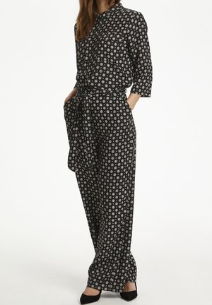 Femme portant une combinaison noire avec des motifs circulaires blancs, manches trois-quarts, taille nouée et talons noirs pointus, debout les mains dans les poches.