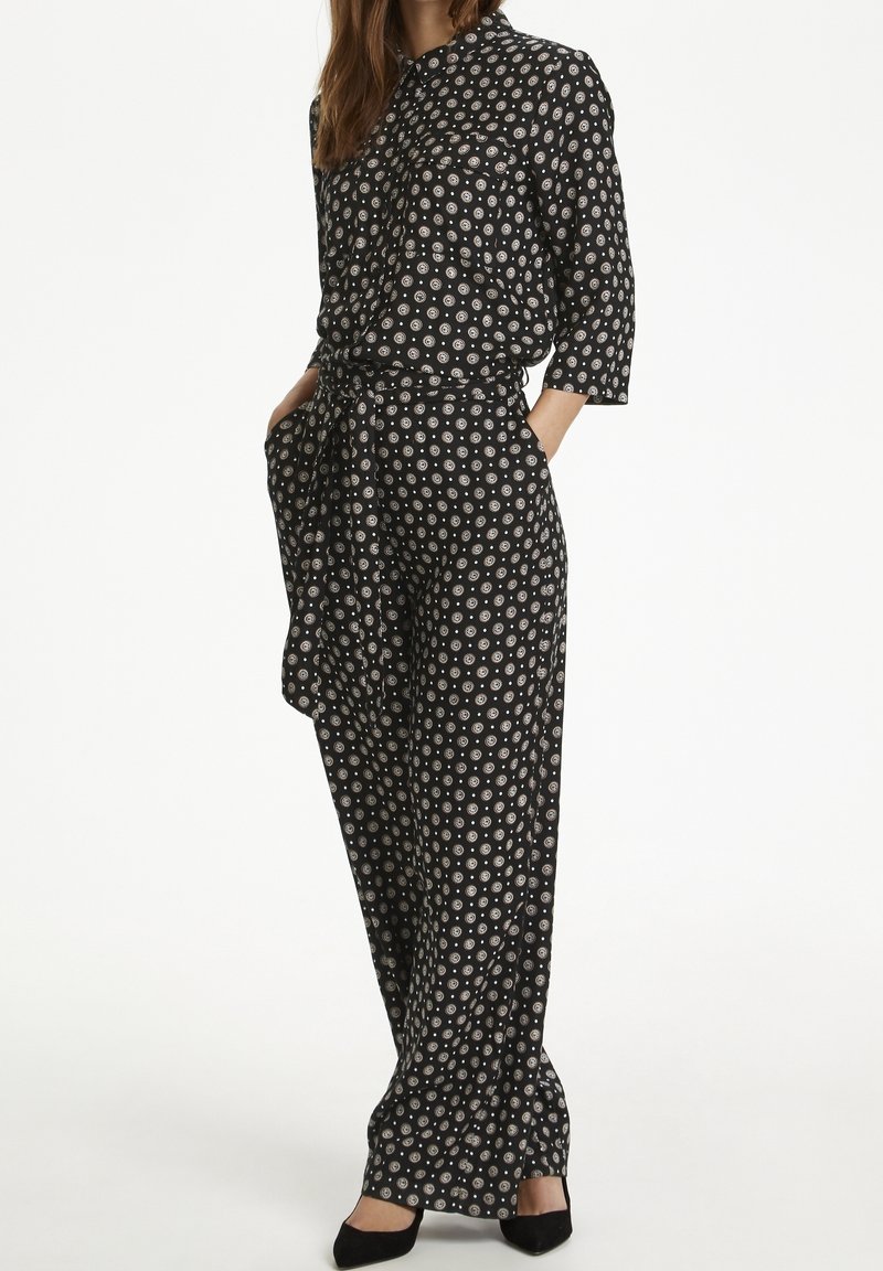 Femme portant une combinaison noire avec des motifs circulaires blancs, manches trois-quarts, taille nouée et talons noirs pointus, debout les mains dans les poches.