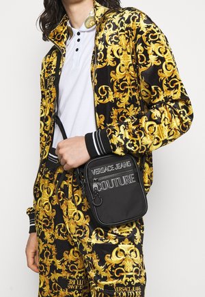 Osoba v černém a zlatém vzorovaném teplákovém kompletu s bílým polokošilí, držící černou crossbody tašku Versace Jeans Couture.