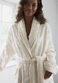 Peignoir crème en tissu texturé avec un motif "self" en relief. Comprend une ceinture, des poches et des manches larges.