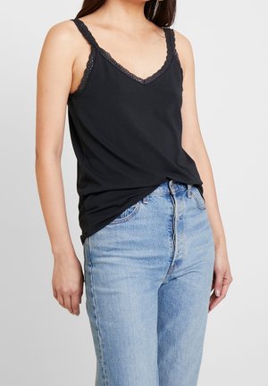 Frau trägt ein ärmelloses schwarzes Top mit Spitzenbesatz, das in eine hellblaue hoch taillierte Jeans gesteckt ist, vor einem einfarbigen Hintergrund.