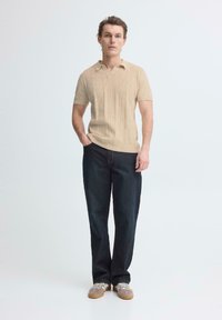 Man staat met handen in de zakken, draagt een beige gebreid T-shirt met korte mouwen, donkere spijkerbroek en beige sneakers tegen een witte achtergrond.