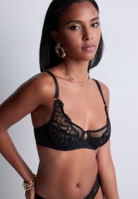 Brassière en dentelle noire avec des panneaux transparents, des motifs floraux et un détail de fermoir doré. Les bretelles réglables et la texture douce augmentent le confort.
