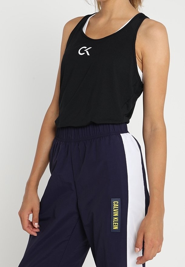 Schwarzes ärmelloses Tanktop mit einem weißen Logo, kombiniert mit navyfarbigen Hosen, die weiße Seitenstreifen und ein gelbes "CALVIN KLEIN"-Label haben.