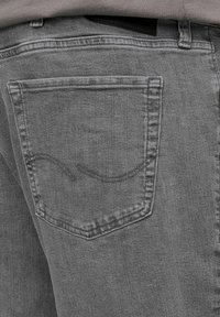 Szare dżinsy z denimu z tylną kieszenią ozdobioną zakrzywionym szwem. Tkanina ma teksturę, jest gładka w dotyku i delikatnie połyskująca.