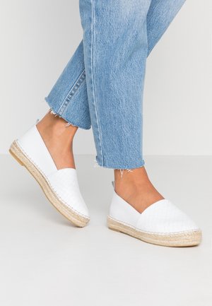 Personne portant un jean court bleu clair et des espadrilles blanches tissées à enfiler avec des semelles en corde sur un fond uni.