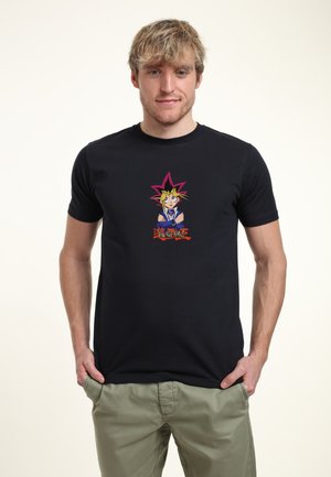Mand med blond hår iført sort Yu-Gi-Oh t-shirt med grafik og grønne bukser, stående med hænderne i lommerne på beige baggrund.