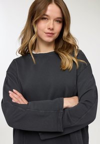 Felpa grigio scuro con scollo rotondo, maniche lunghe e dettagli di cuciture visibili. Il tessuto appare morbido e crea una vestibilità comoda.