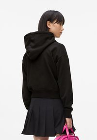 Giovane donna con capelli neri lisci sta in posizione laterale indossando un hoodie nero e una gonna plissettata nera, tenendo una piccola borsa rosa al suo fianco.