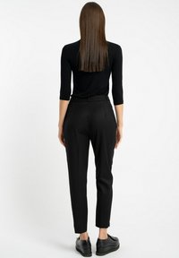 Czarne dopasowane spodnie o tapered kroju i subtelnymi zakładkami, zestawione z czarną bluzką z długim rękawem, widziane z tyłu. W komplecie czarne obuwie.