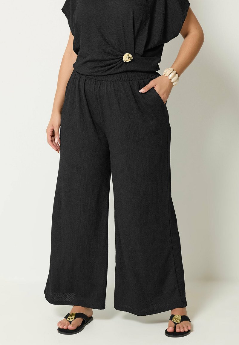 Woman wearing black wide-leg pants, black top with gold pin, shell bracelet, and black sandals with gold detail, standing against a light background.