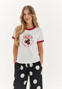 Bílé tričko s krátkým rukávem a červeným lemem, secherry grafikou a textem. Černé kalhoty s velkými bílými puntíky. Volný střih.