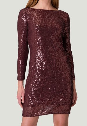 Frau trägt ein eng anliegendes, langärmeliges, knielanges Kleid, bedeckt mit glänzenden, dunkelvioletten Pailletten, vor einfachem Hintergrund.