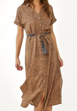 Kobieta ubrana w beżową sukienkę midi w czarne plamki, z krótkimi rękawami, z przodu zapinaną na guziki i przewiązaną czarno-białym paskiem w grochy.