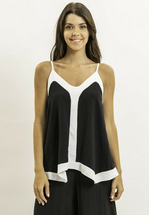 Jeune femme souriante, portant un haut sans manches noir et blanc à fines bretelles et un pantalon noir ample, debout devant un fond clair.