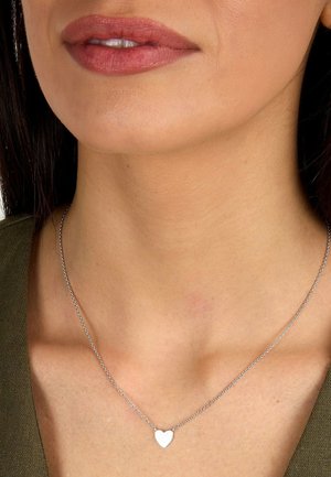 Nærbillede af en persons hals og læber, der bærer en sølvfarvet hjerteformet vedhæng og en olivengrøn top.