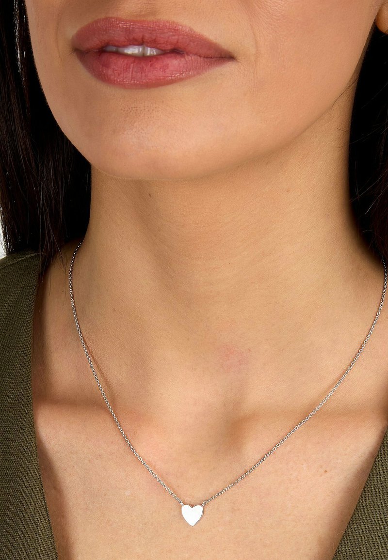 Nærbillede af en persons hals og læber, der bærer en sølvfarvet hjerteformet vedhæng og en olivengrøn top.