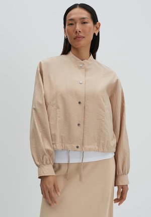 Donna con capelli neri lisci che indossa una giacca beige con bottoni, polsini elastici e orlo con coulisse, abbinata a una gonna coordinata e a una maglietta bianca.