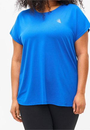 Žena nosící volné krátké modré tričko s malým bílým logem na levé straně hrudníku, doplněné černými kalhotami.