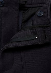 Detail námorníckeho saku ukazujúci textúrovanú tkaninu, gombíky a zips na ľavej strane s prešitými švami a kontrastnou podšívkou.