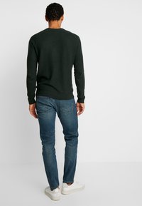 Dunkelgrüner Strickpullover mit langen Ärmeln, kombiniert mit blauen Jeans. Weiße Sneakers runden das lässige Outfit ab.