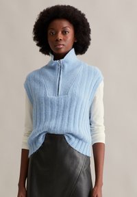 Himmelblaues, geripptes Strickoberteil mit Reißverschlusskragen, kurzen Ärmeln und seitlichen Schlitzen, kombiniert mit einem enganliegenden weißen Langarmshirt und einem schwarzen Lederrock.