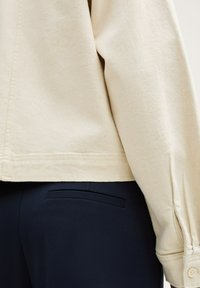 Hanorac din bumbac crem, cu croială lejeră, având manșete cu nasturi și textură fină, asortat cu pantaloni tăiați negri închisi, având un buzunar pe spate.