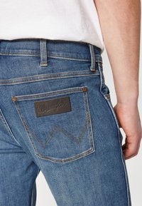 Jeans din denim albastru, cu un patch din piele și cusături contrastante de culoare portocalie. Buzunarul din spate are un model de cusătură în zigzag.
