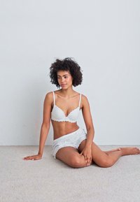 Brassière en dentelle blanche et short assorti avec des bords festonnés et une texture douce, assis sur un tapis clair contre un mur neutre.