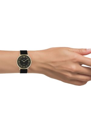 Montre noire avec accents dorés, cadran rond, face texturée, chiffres romains, et bracelet en nylon, présentée sur une main sur fond blanc.