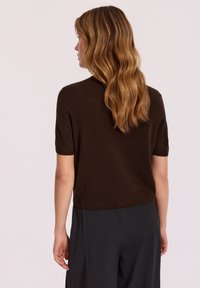 Brązowy sweter dzianinowy z krótkim rękawem o luźnym kroju i ściągaczem na dolnej krawędzi. Miękka tekstura. Połączony z czarnymi spodniami o szerokich nogawkach, widok z tyłu.