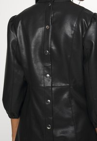 Veste en similicuir noir avec manches bouffantes et boutons-pression argentés courant le long de la couture centrale arrière.