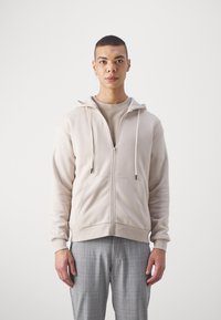 Sudadera beige clara con cremallera y capucha ajustable, bolsillos frontales y puños de canalé, combinada con pantalones de cuadros grises. Textura suave.