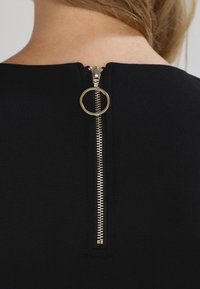 Černá žebrovaná látka se středovým zipem na zadní straně, vybavená oblíbeným kroužkem a kovovými zuby. Minimalistický a funkční design.