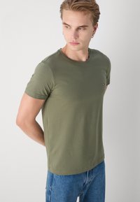 Olivenfarbendes, kurzärmeliges T-Shirt aus weichem Stoff. Runder Ausschnitt und kleines Logodetail auf der linken Seite. Lässige Passform.