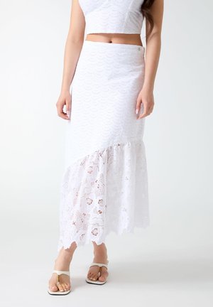 Frau trägt ein weißes gehäkeltes zweiteiliges Outfit mit Crop-Top und asymmetrischem Spitzenrock, kombiniert mit weißen Sandalen.