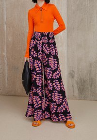 Haut orange à manches longues avec un col zippé, associé à un pantalon noir à jambes larges ornés de motifs de feuilles roses, et des sandales orange à enfiler. Sac noir à la main.