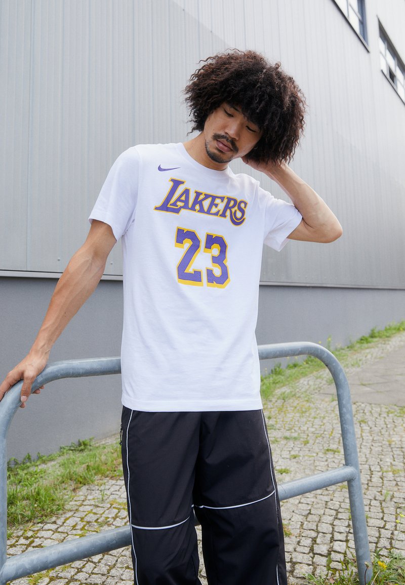 Hvid bomulds t-shirt med lilla og gule "Lakers" grafik og nummer 23. Inkluderer Nike-logo. Kombineret med sorte træningsbukser med hvide detaljer.