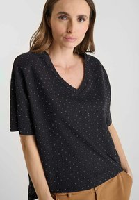 Camiseta negra de cuello en V con mangas cortas, con un patrón de manchas en blanco. Tejido suave con un ajuste relajado y costuras visibles.