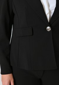 Zwarte blazer van gladde stof, voorzien van een enkele zilveren knop, een opstaande kraag en een zijzakdetail.