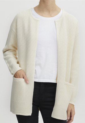 Cardigan texturé, couleur crème, avec une coupe ample, un devant ouvert et deux poches avant. Porté par-dessus un t-shirt blanc et un pantalon noir.