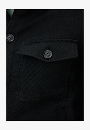 Zwarte geweven stoffen jas met een borstzak met knoopsluiting. De textuur is glad met een lichte glans.