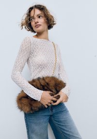 Weißes Spitzenshirt mit langen Ärmeln und floralem Muster, kombiniert mit einer braunen Fake-Fur-Clutch-Tasche mit einer goldenen Kettenriemen. Blaue Denim-Jeans runden den Look ab.